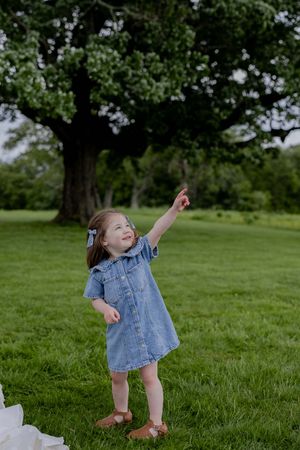 Child in denim dress pointing up at sky while playing on grassy field.