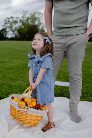 Child in denim overalls carrying a yellow wicker basket filled with fruit at a picnic.