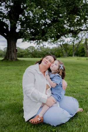 Parent and child sitting together on green grass under a large tree in a peaceful park setting.