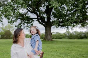 Parent holding child in denim dress while standing in green field under large tree.