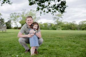 Parent and small child in denim dress embrace while kneeling on grassy field during outdoor photo session.