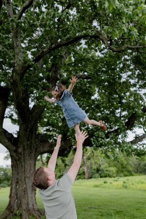 Playful outdoor scene of a child being tossed in the air near a large tree in a grassy field.