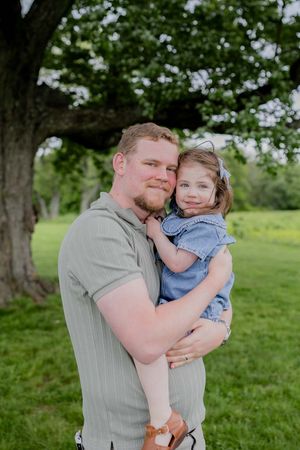 A parent and child share a tender moment while posing under a shady tree in a green field.