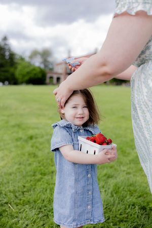 A denim-dressed child holds a bowl of strawberries while standing on grass with a house in the background.