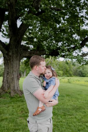 A sweet outdoor moment captured under a majestic tree in a lush green setting.