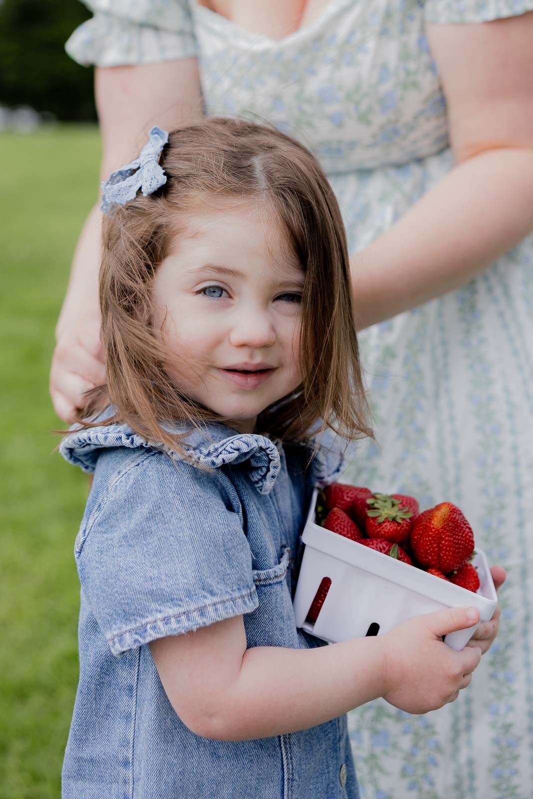 A small child in a denim dress holding a container of fresh red strawberries outdoors.
