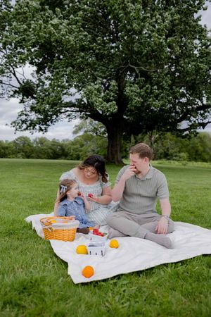 Family enjoying a summer picnic under a large tree with fruits and snacks on a white blanket.