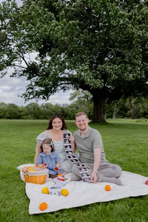 A family enjoys a sunny picnic on a blanket under a large tree with oranges and a basket in a grassy field.
