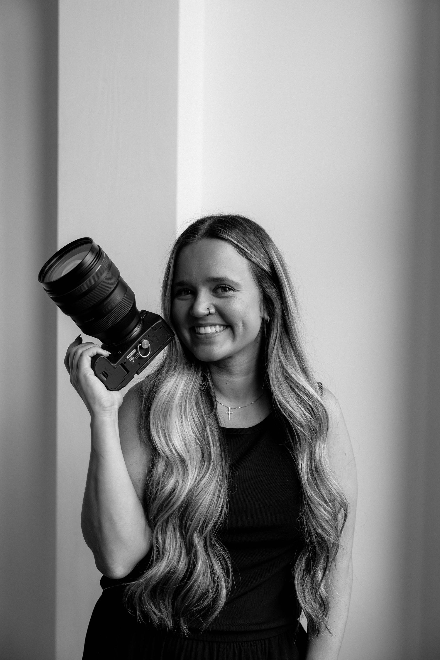 A photographer in a black top holding a professional camera smiles against a white background.