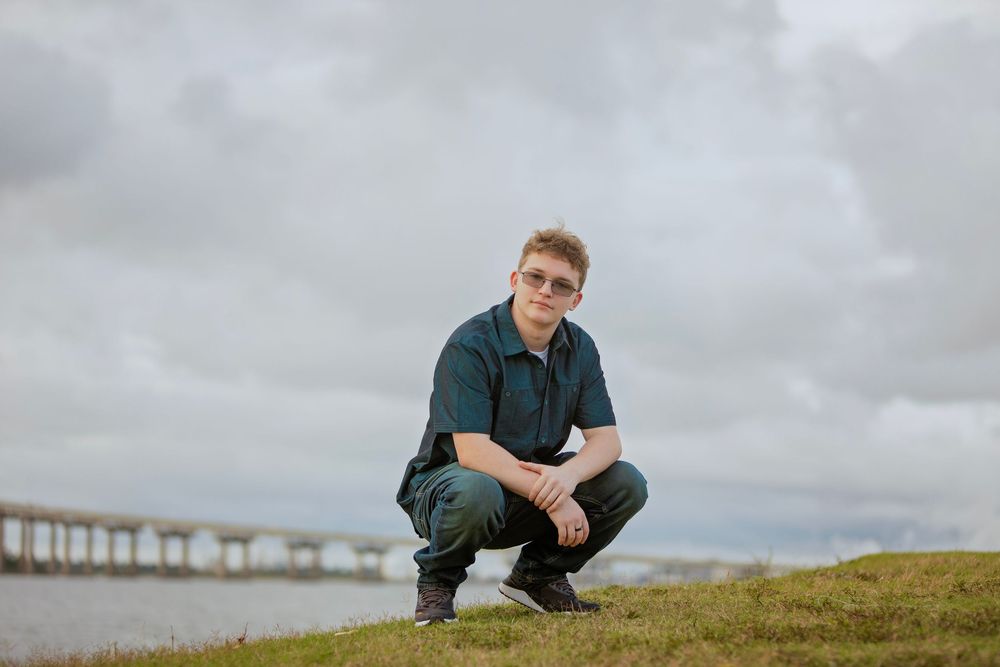 A person crouches on a grassy hill with a long bridge stretching across the water in the background.