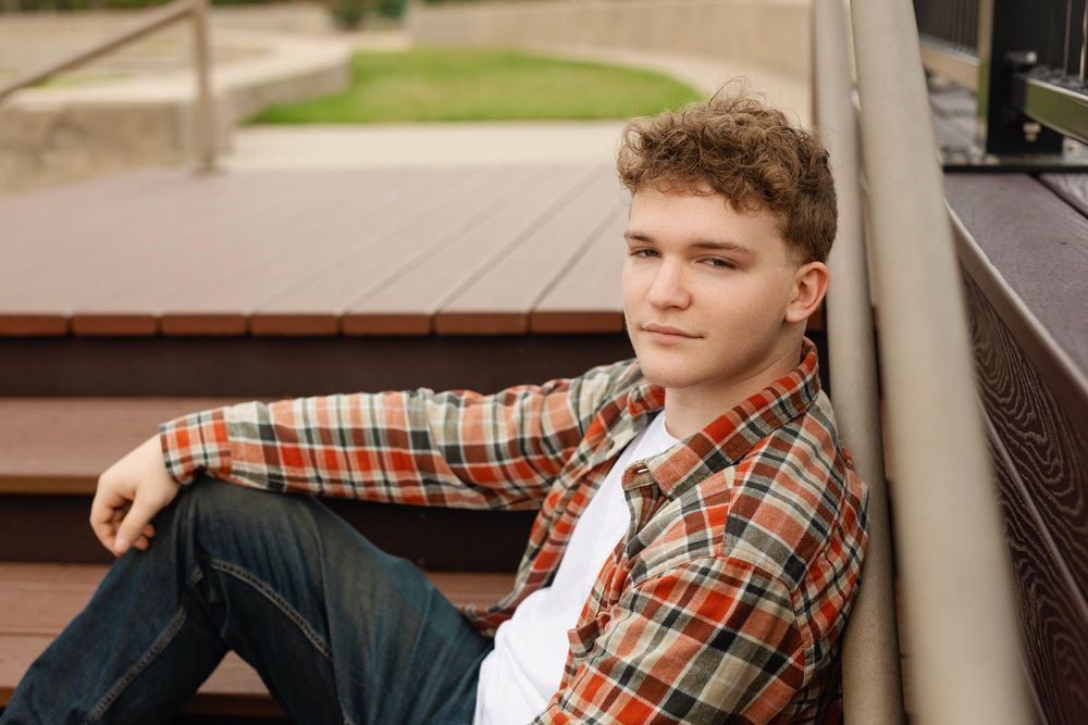 Young person in plaid shirt and jeans lounging casually on wooden steps outdoors.