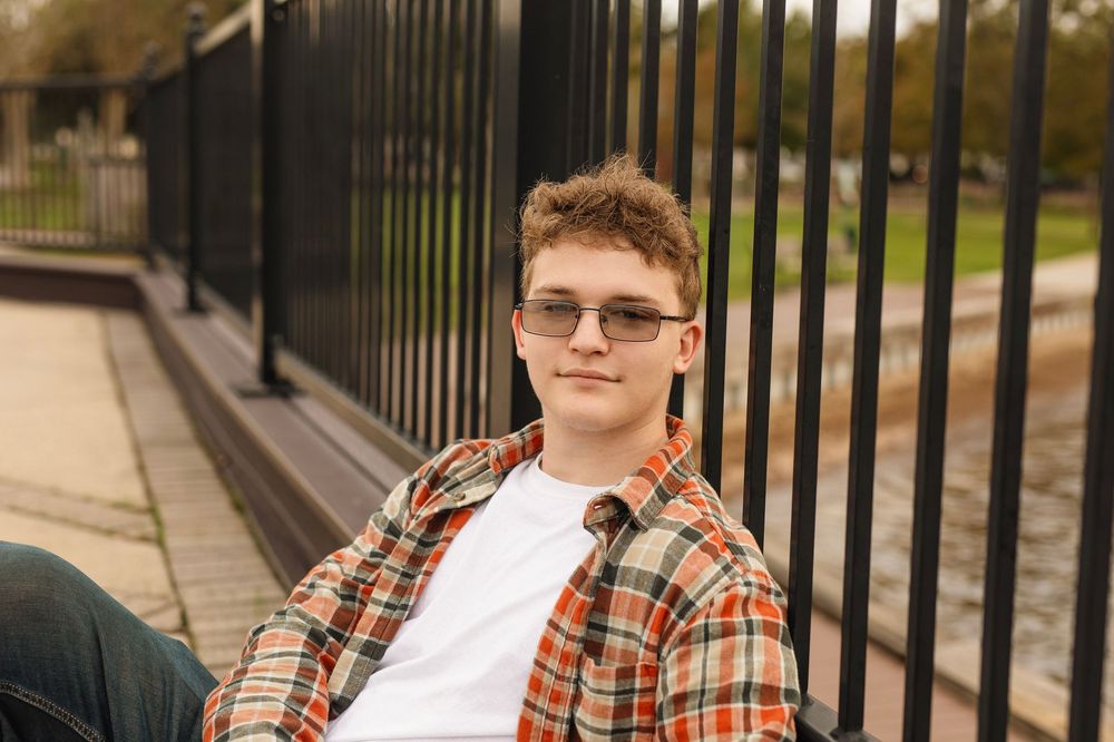 Stylish person in plaid shirt and sunglasses sits casually against metal railing on sunny day outdoors.