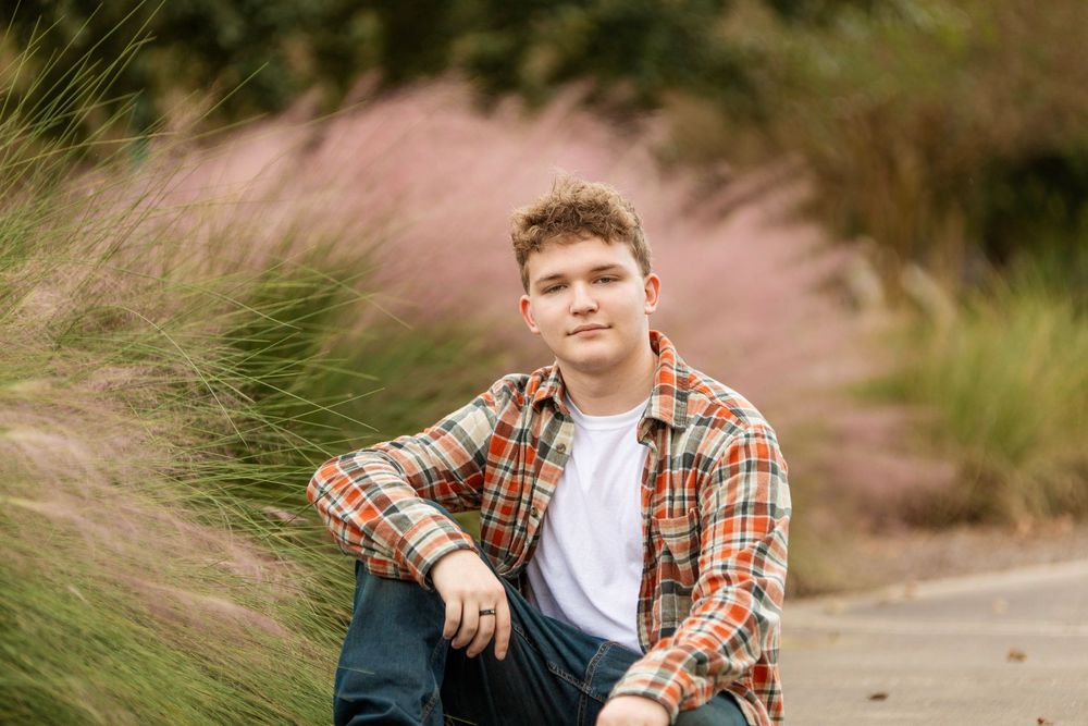 A casual outdoor portrait in a plaid flannel shirt seated among tall ornamental grasses during golden hour.