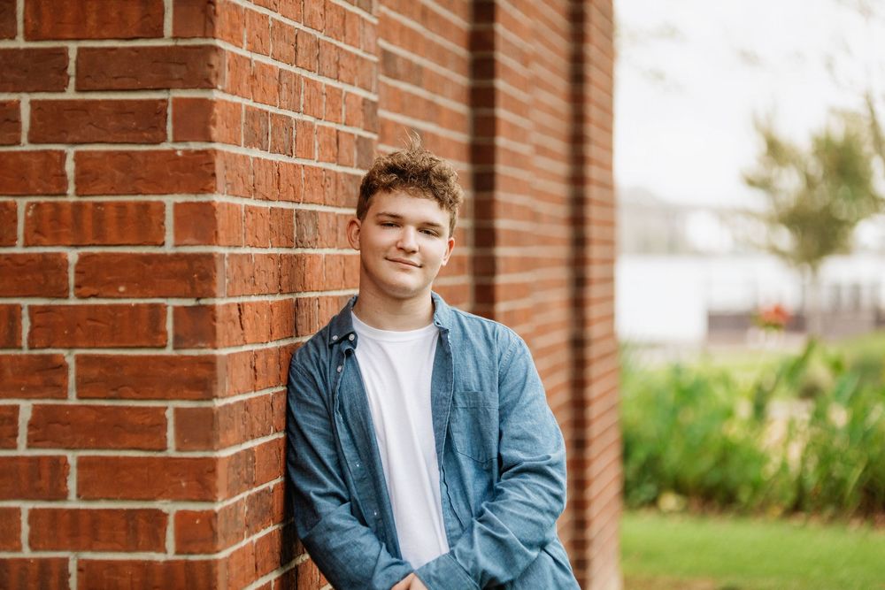 Someone wearing a denim jacket and white t-shirt leans casually against a red brick wall outdoors in natural lighting.