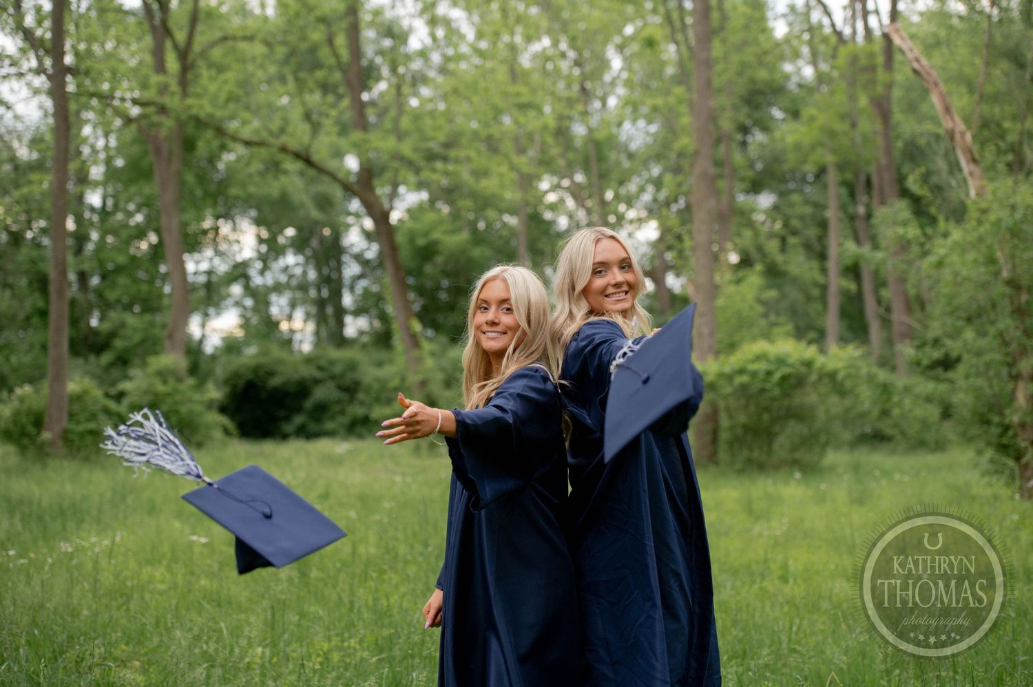 Graduates in navy blue caps and gowns toss their graduation caps while celebrating in a green field.