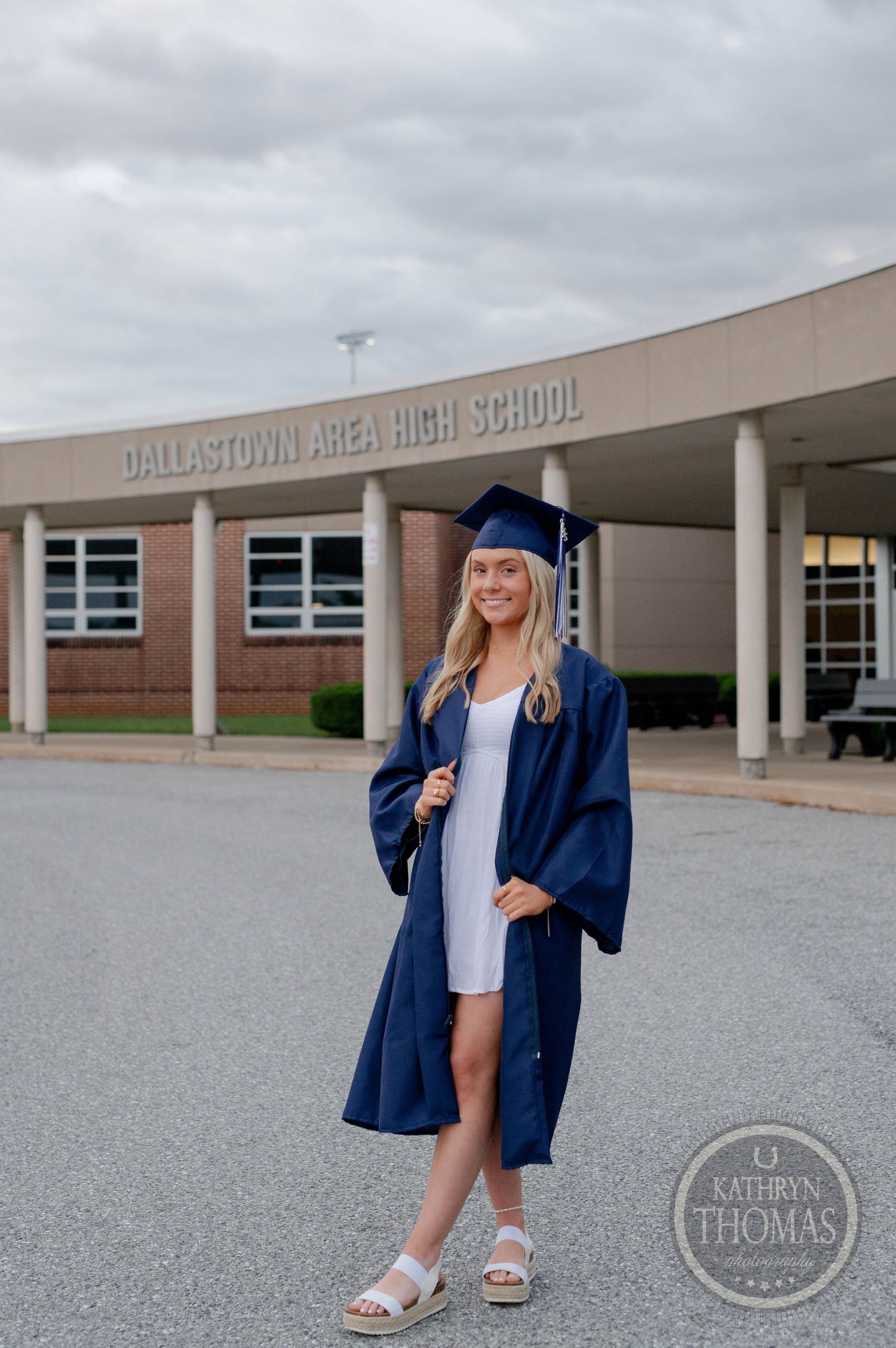 Graduate wearing navy blue cap and gown poses in front of Dallastown Area High School on graduation day.