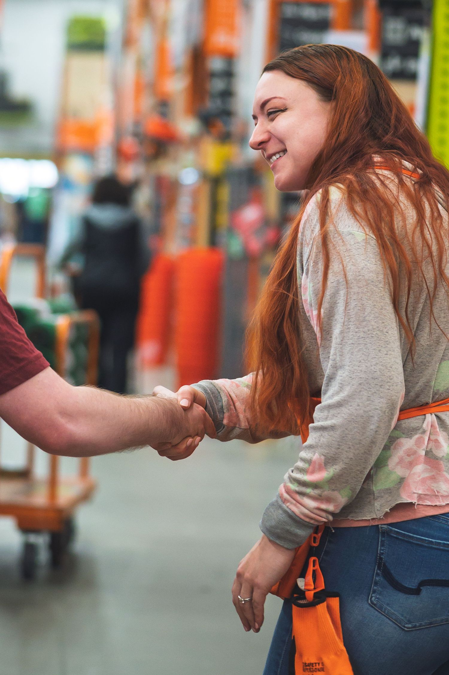 An employee in a gray sweater shakes hands with a customer while carrying an orange shopping bag in a retail store.