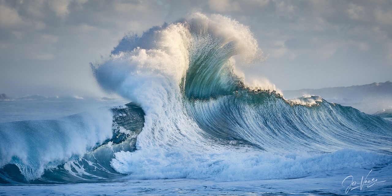 Powerful ocean waves with turquoise water break and curl dramatically against a cloudy sky in a panoramic seascape.