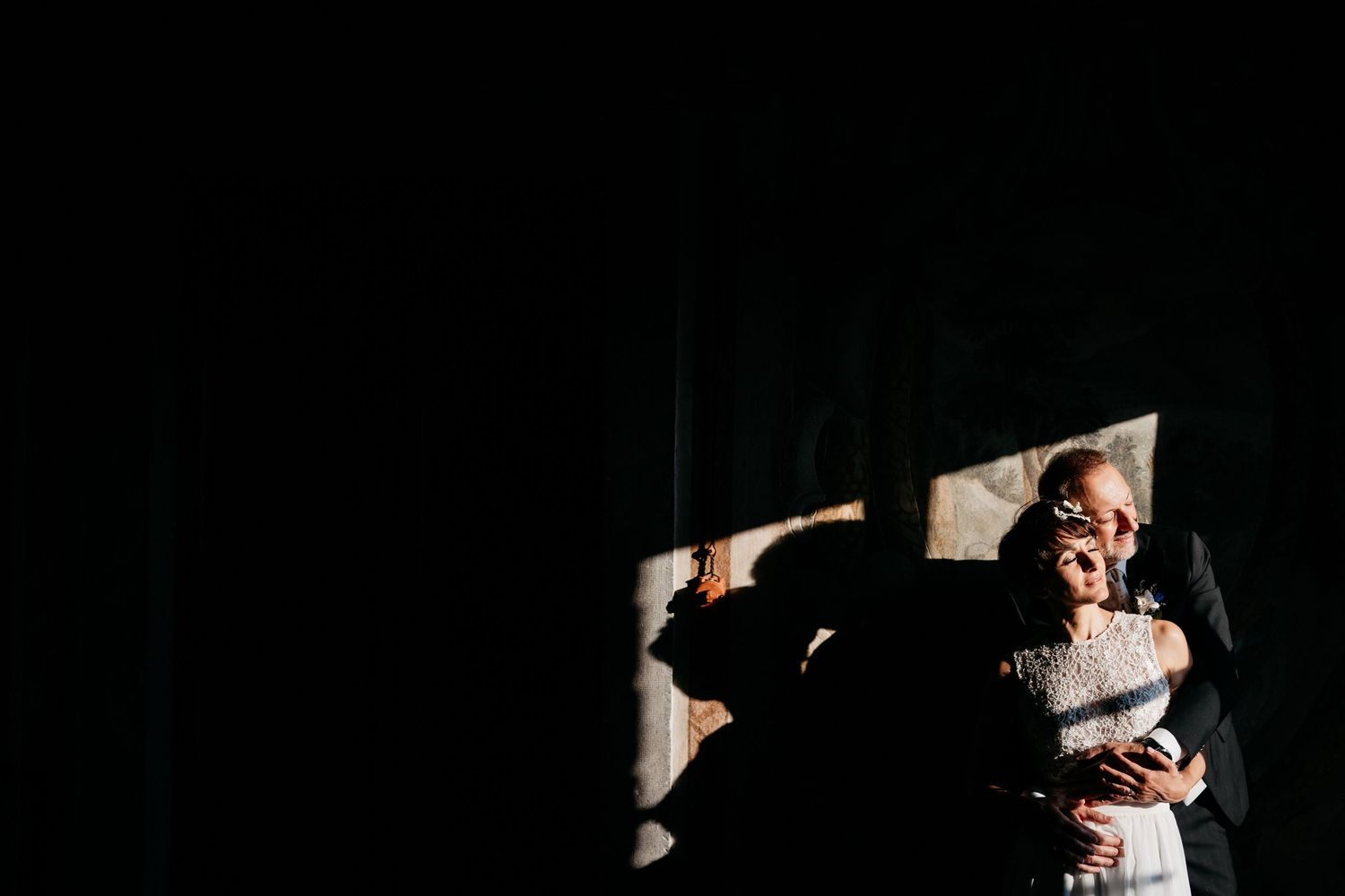 Couple embraces in dramatic dark setting with diagonal light beams creating atmospheric shadows and highlights.