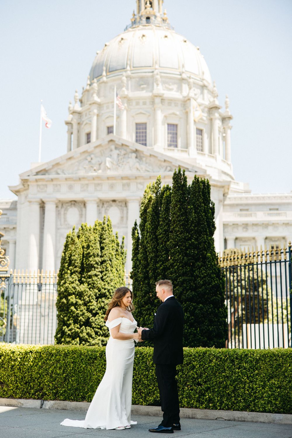Couple exchanging vows outside San Francisco City Hall, bride in white gown, groom in black suit, sunny day.