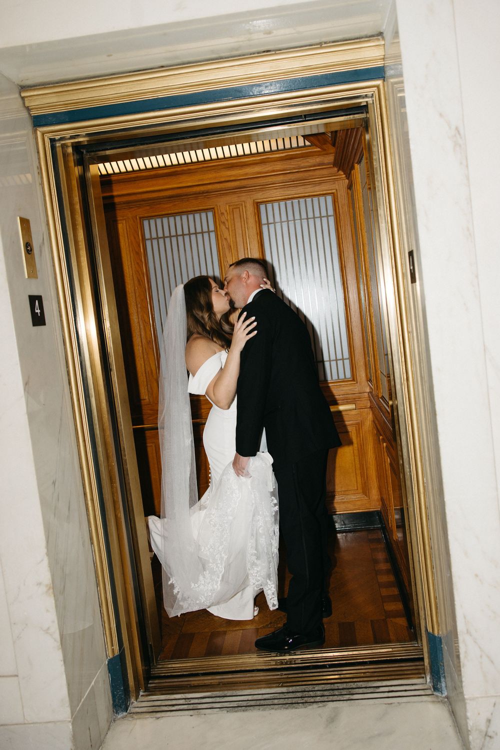 Bride and groom sharing a romantic kiss inside an ornate gold elevator with wooden doors on their wedding day.