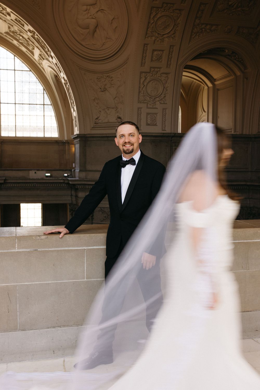 Bride in white gown with flowing veil and groom in black tuxedo posing inside San Francisco City Hall.