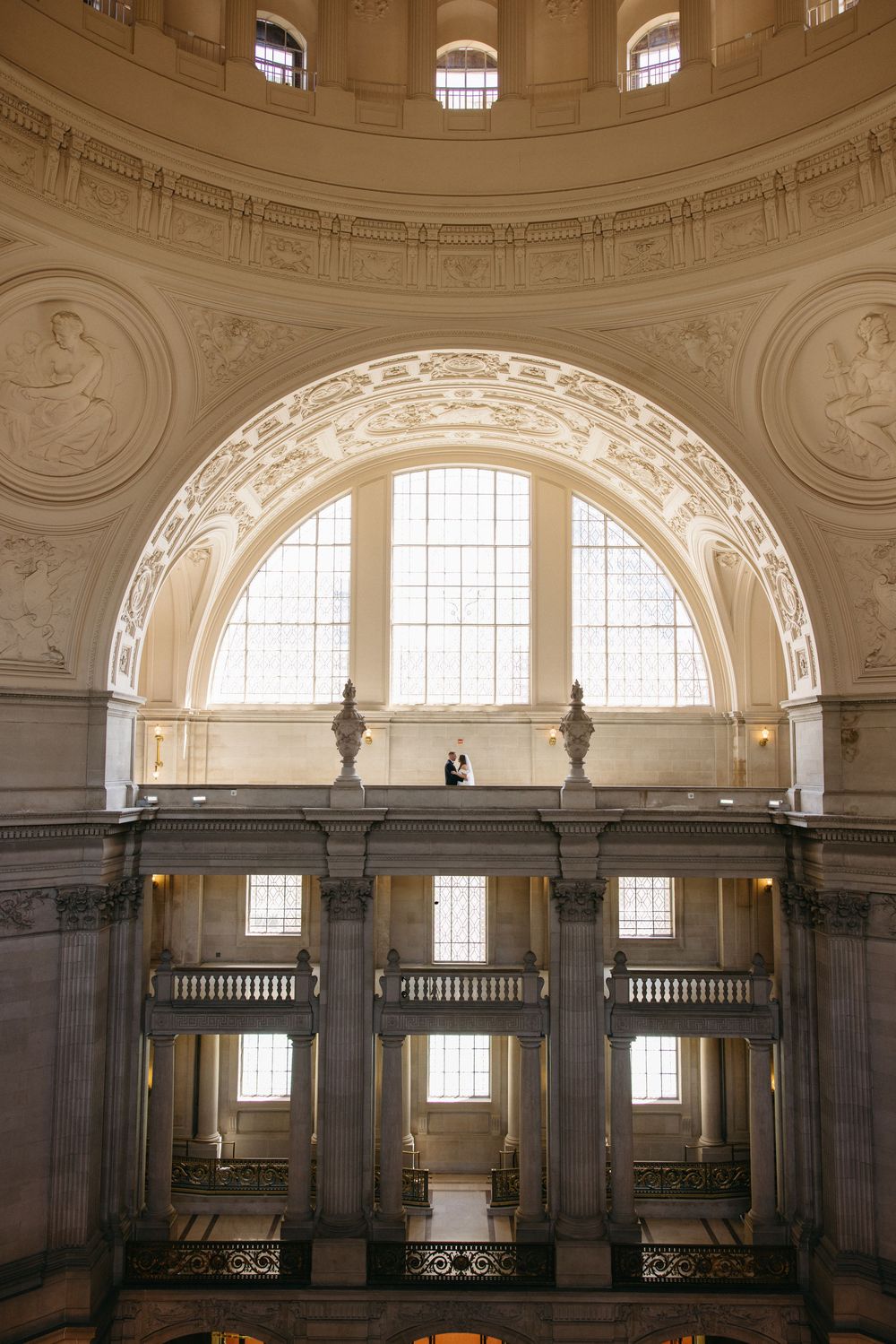 Couple stands on grand balcony inside San Francisco City Hall rotunda with ornate arched windows and classical architecture.