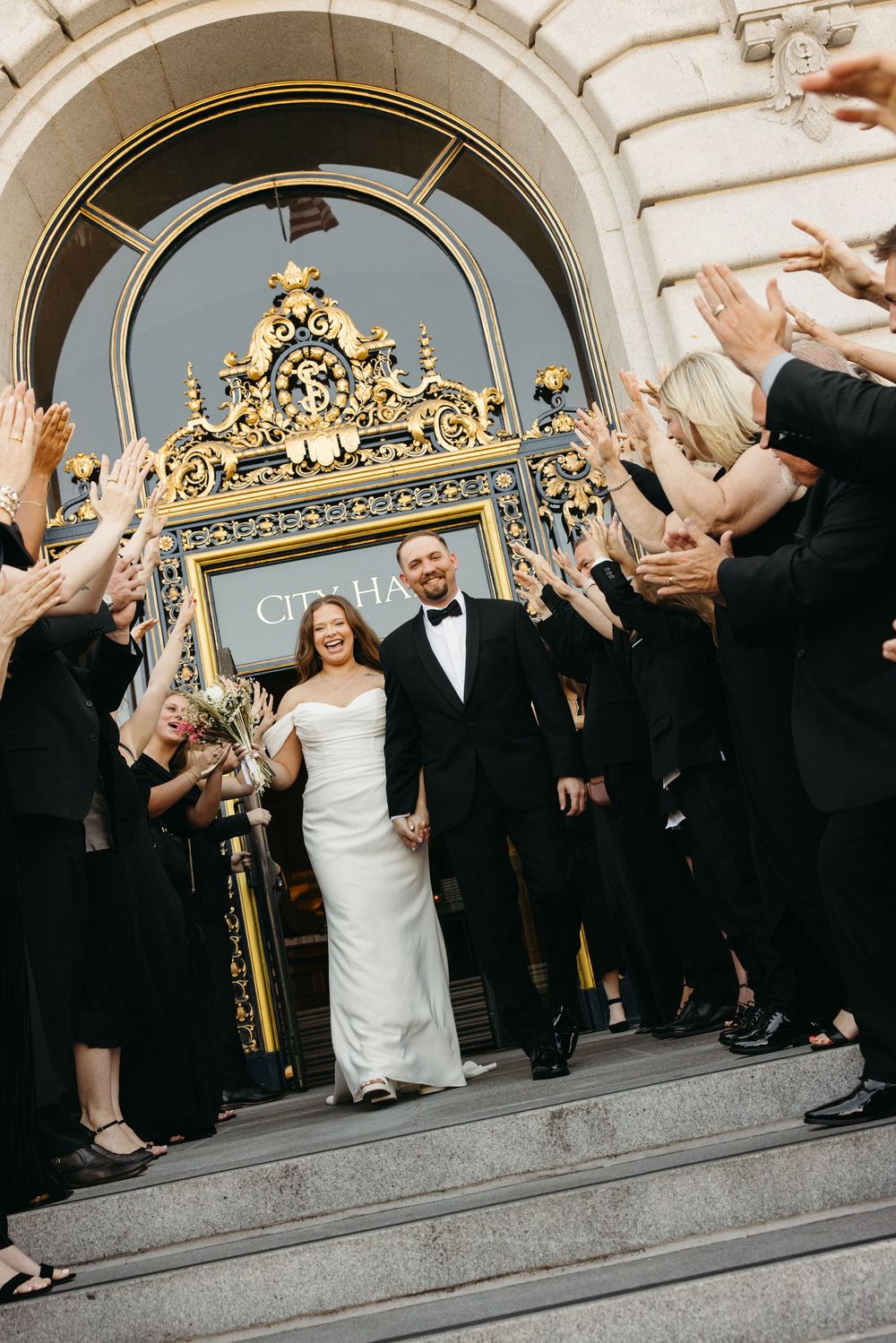 Bride and groom exit San Francisco City Hall in wedding attire as guests in black celebrate on the steps.