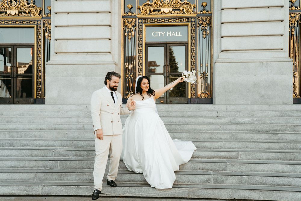 A couple in elegant white wedding attire poses on grand marble steps of a historic city hall building.