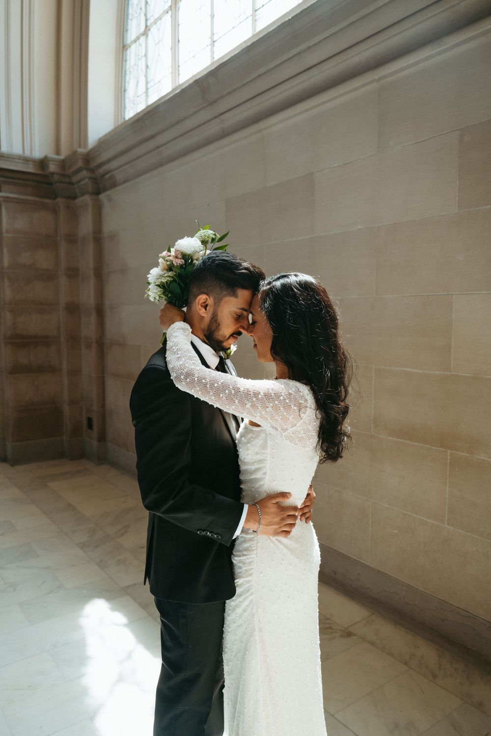 A romantic wedding couple embraces in a bright sunlit room with stone walls and tall windows.