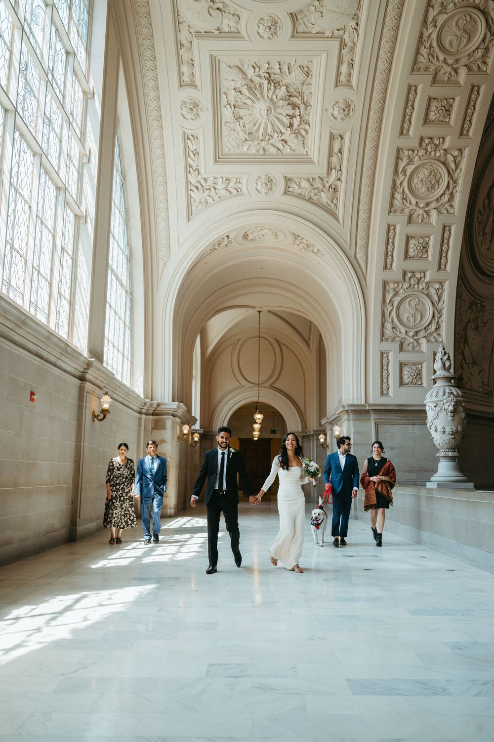 Elegant couple and guests walk through ornate marble hallway with decorative ceiling and large windows in City Hall building.