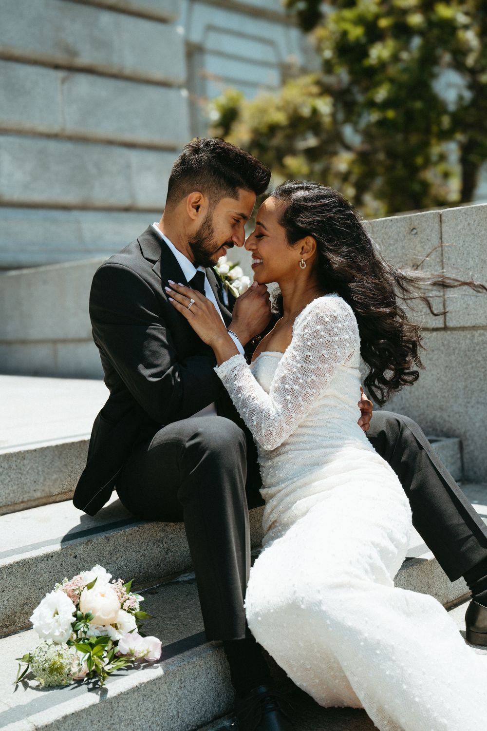 Newlywed couple shares an intimate moment on stone steps with white bouquet laying beside them on a sunny day.