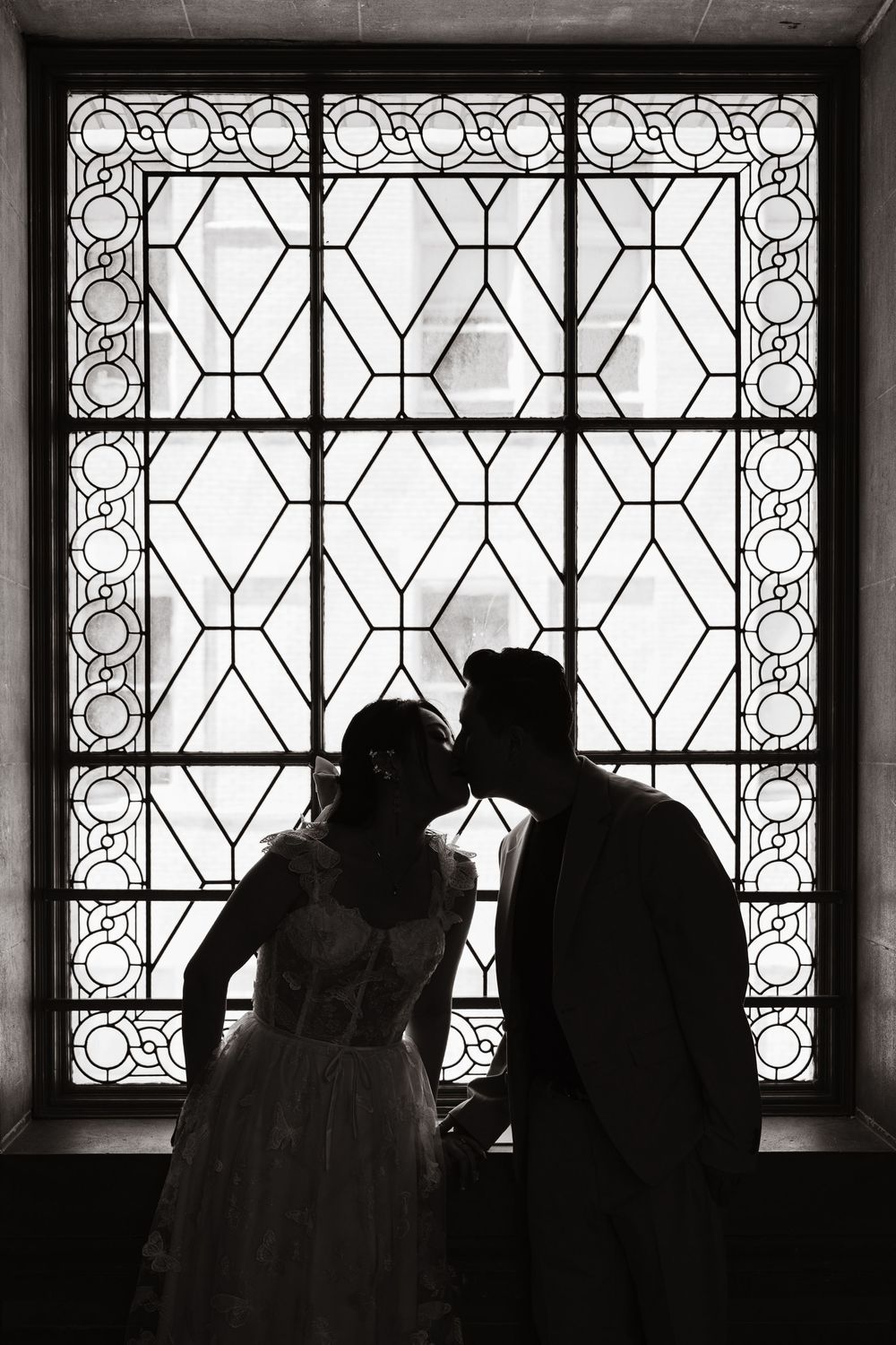 Silhouette of a romantic couple standing in front of an ornate stained glass window with geometric patterns.