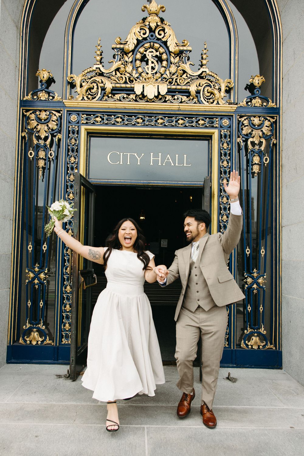 Joyful newlyweds celebrate outside San Francisco City Hall, bride in white dress holding bouquet, groom in tan suit, waving.