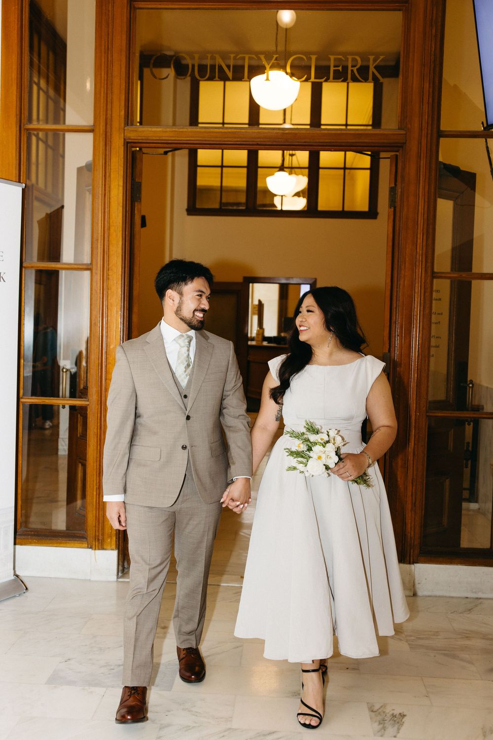 Newlywed couple holding hands and smiling outside County Clerk office, bride in white dress with bouquet, groom in tan suit.