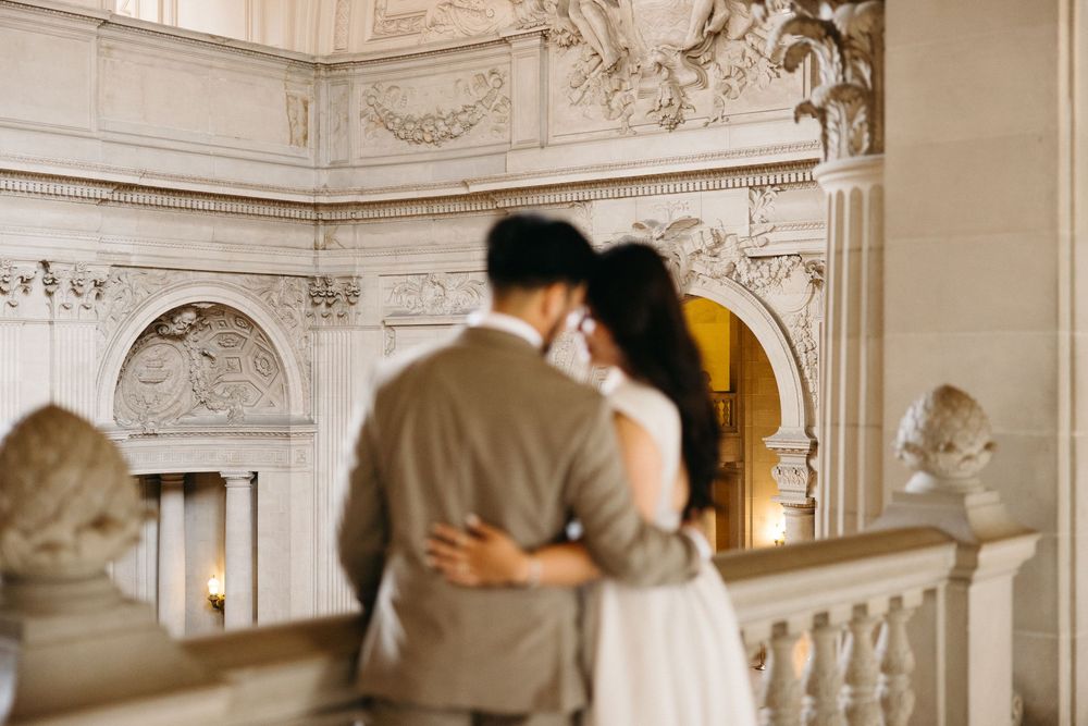 Couple embracing on elegant marble balcony at San Francisco City Hall, surrounded by ornate classical architecture.