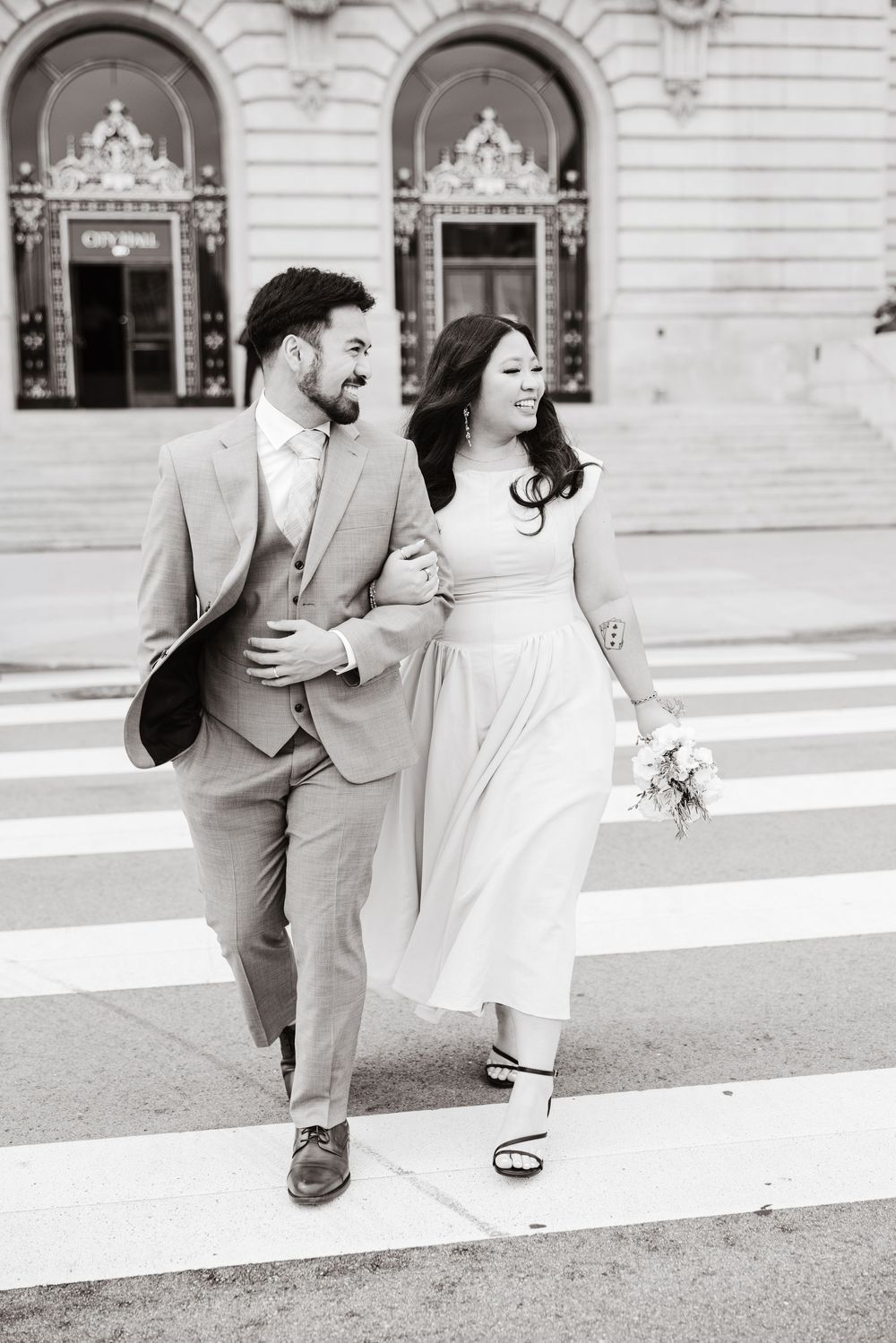 Black and white photo of a couple in wedding attire walking arm in arm across a crosswalk outside City Hall.