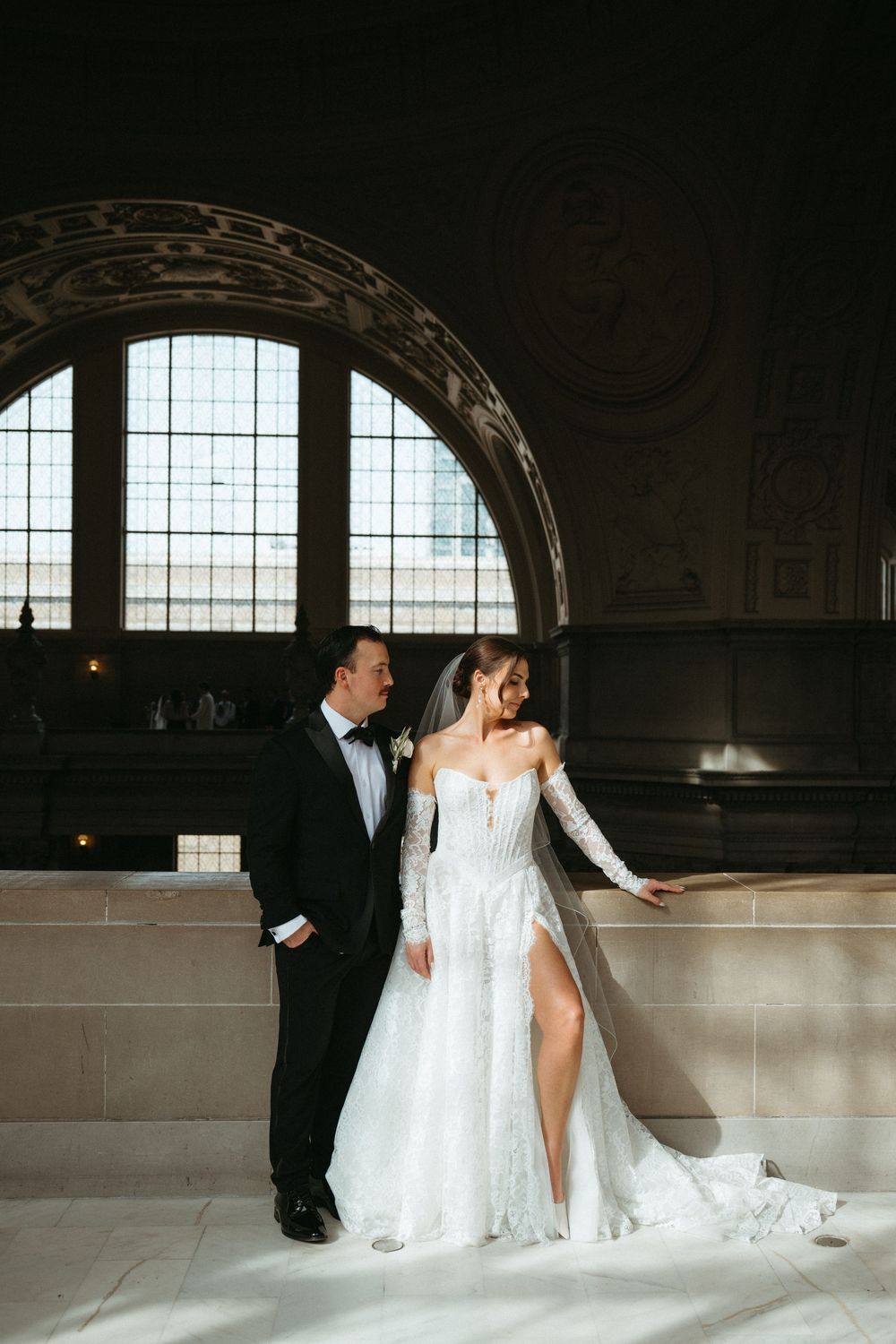 A romantic wedding couple poses in an elegant venue with arched windows and natural light streaming in.