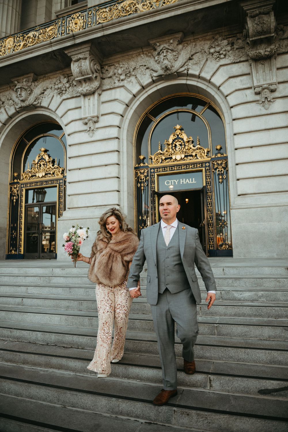A couple descends elegant stone steps outside an ornate building with gilded doorways and decorative architectural details.