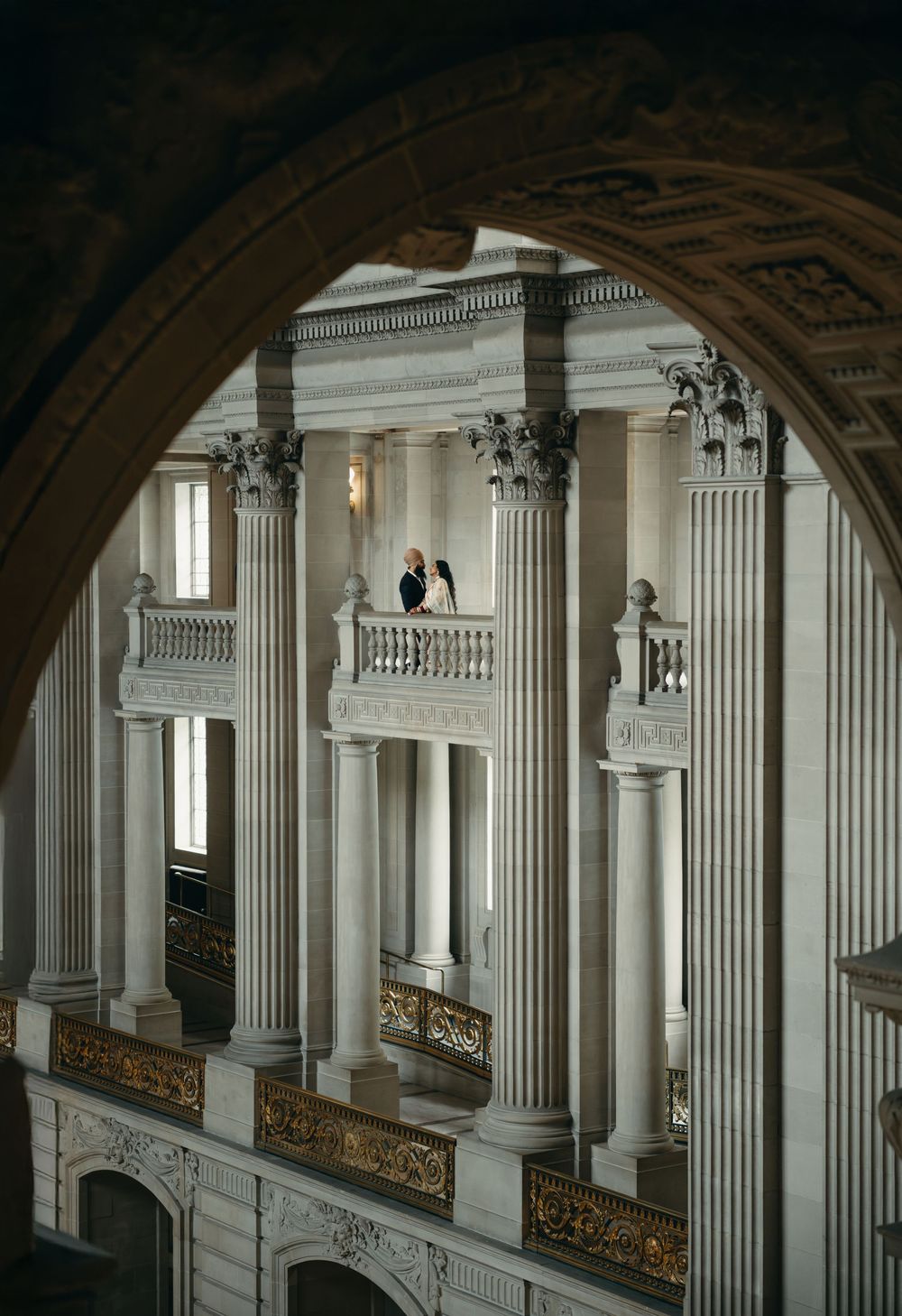 Interior view of ornate neoclassical architecture with white columns and decorative balconies framed through an archway.