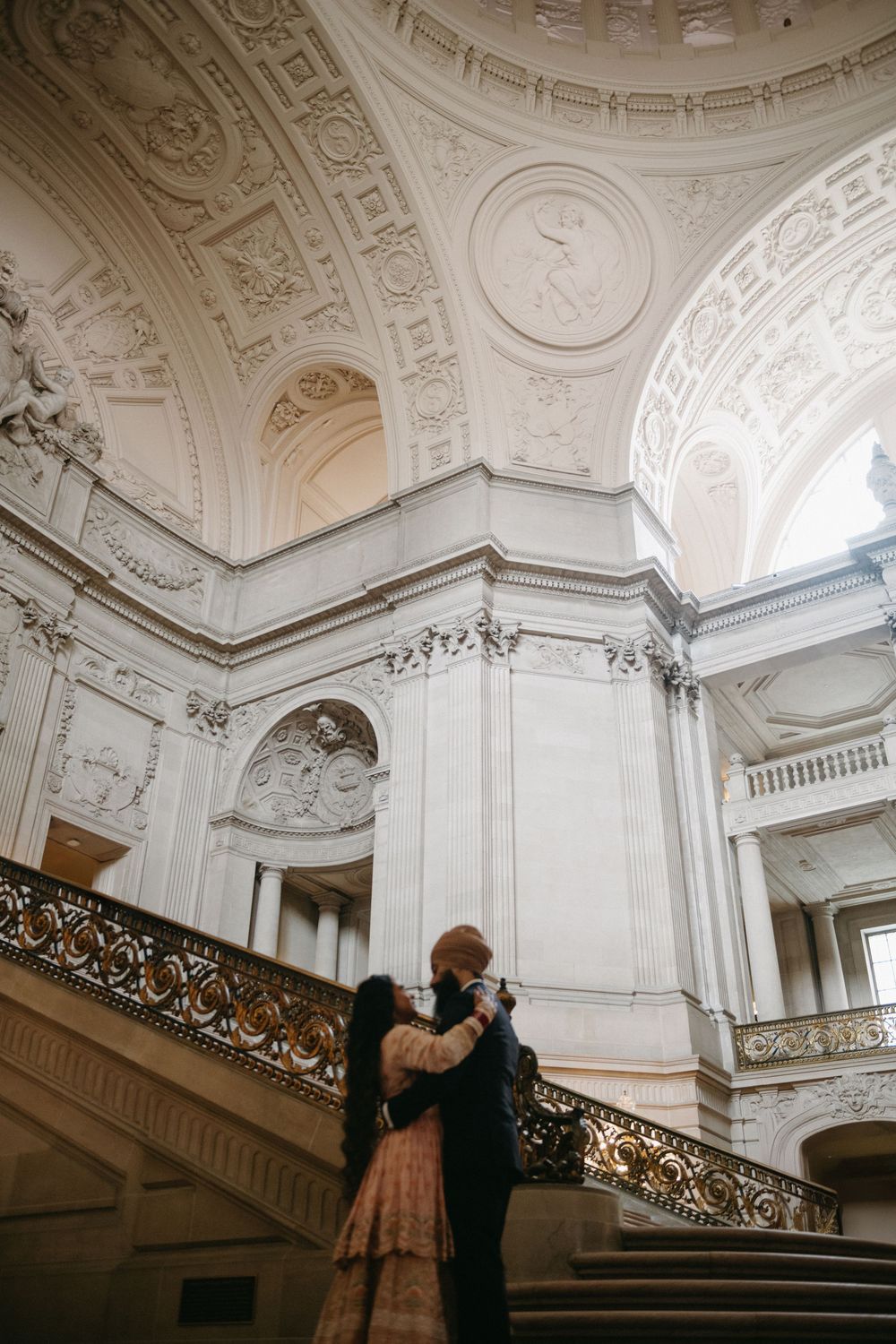 A couple standing on an ornate marble staircase inside a grand historic building with decorative architectural details.