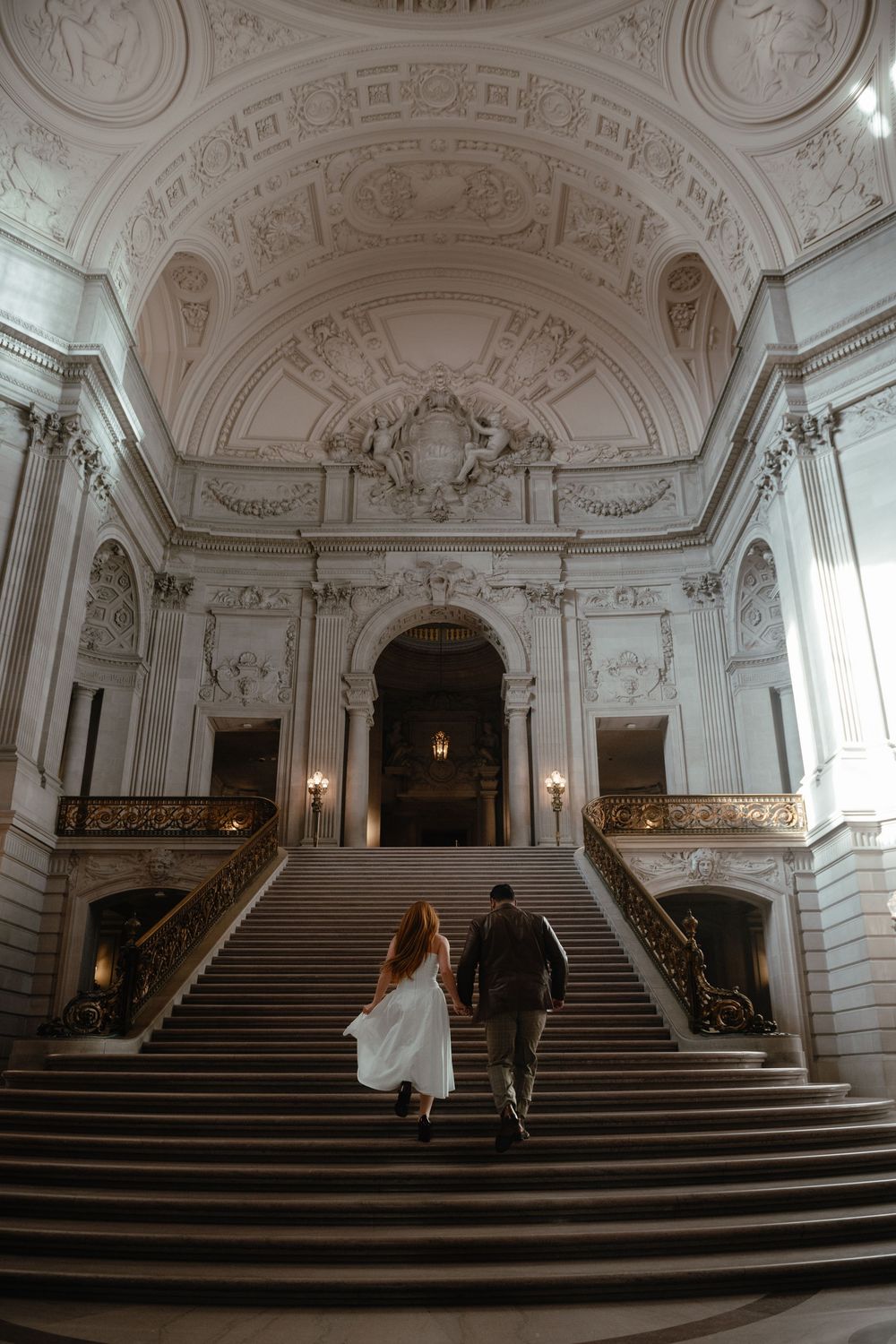 Two people ascend an ornate marble staircase in a grand historic building with elaborate baroque architectural details.