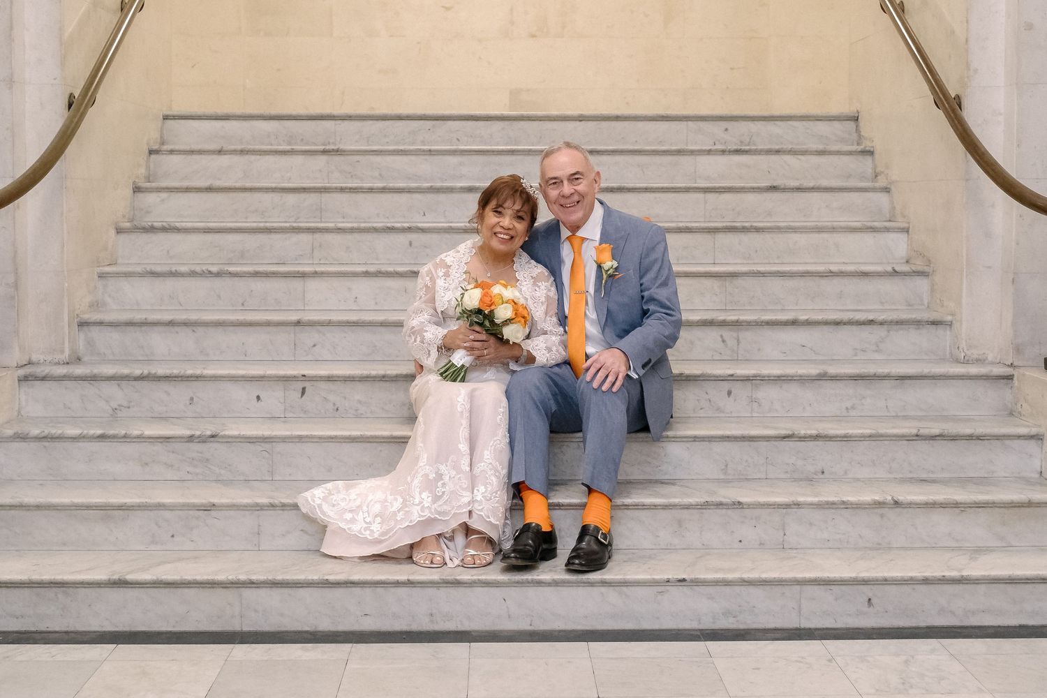 A wedding couple sits intimately together on ornate marble stairs with a white bouquet in a romantic moment.