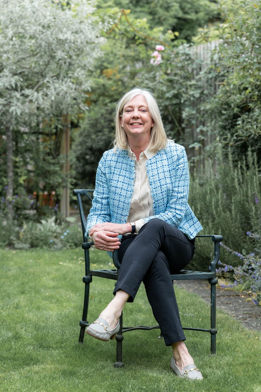 A person in a blue checkered blazer relaxes outdoors in a garden chair on a sunny day with blooming flowers in background.