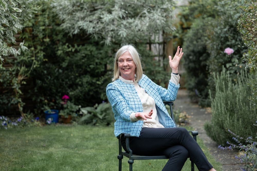 Someone wearing a blue checkered shirt sits in a garden chair giving a peace sign while smiling in a lush backyard setting.
