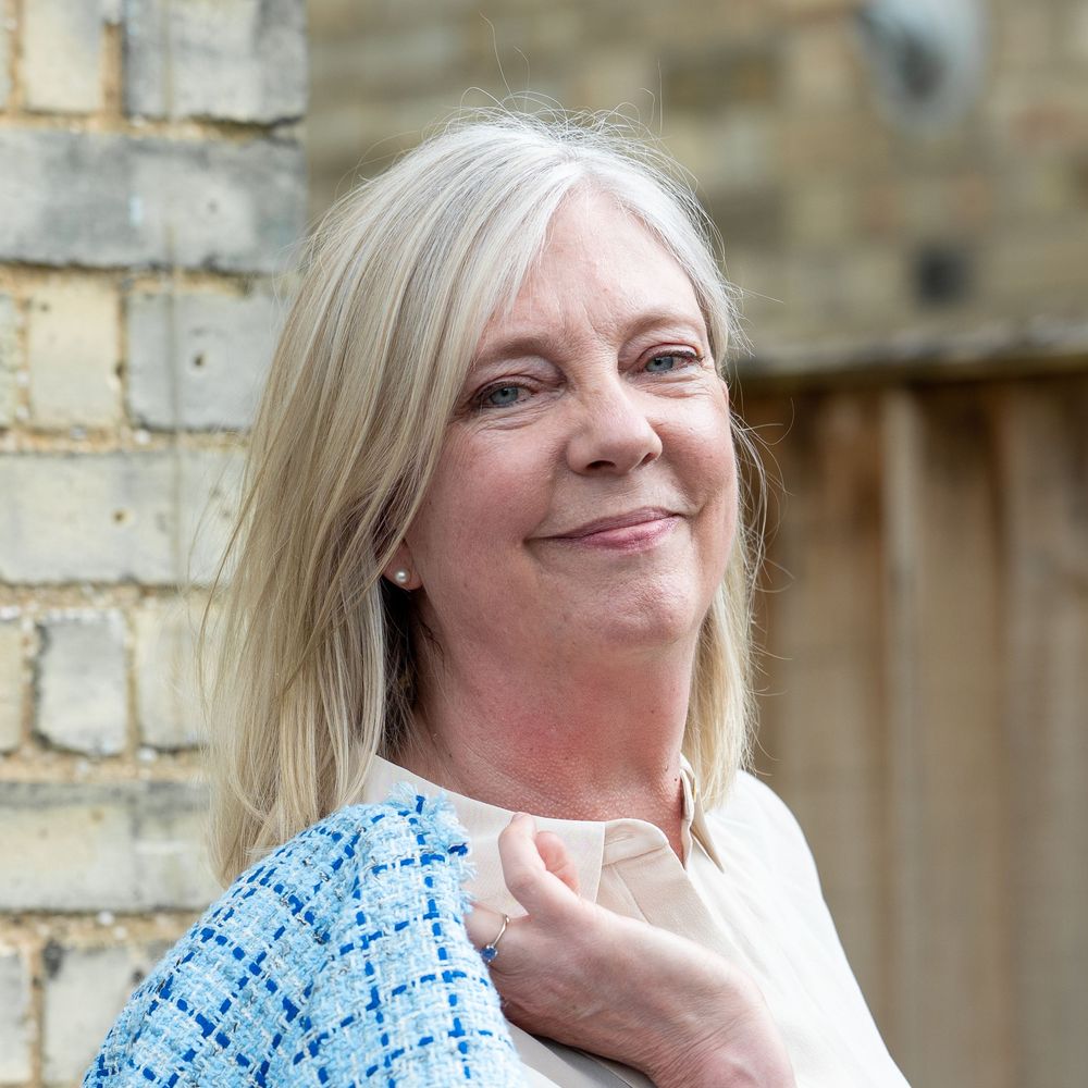A middle-aged woman with blonde hair smiling while leaning against a brick wall wearing a blue patterned top.