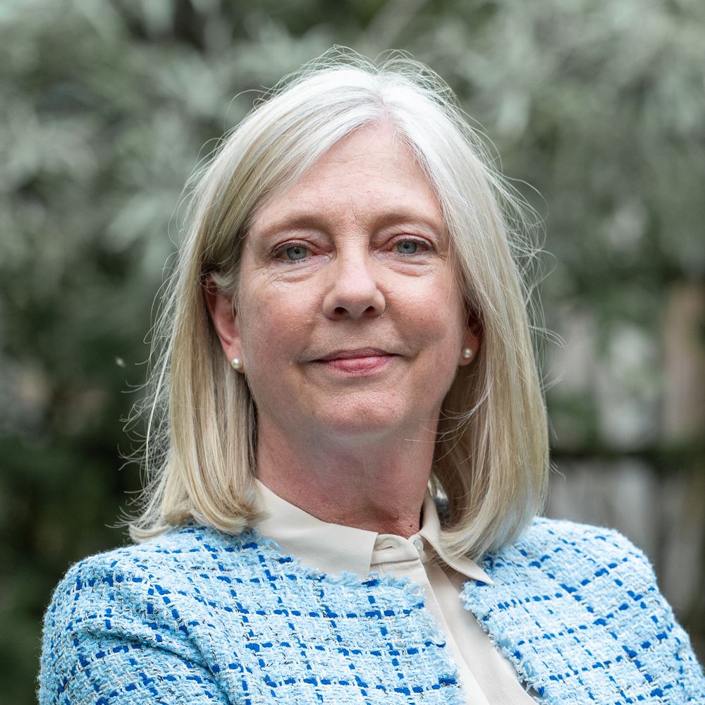 Professional headshot of someone wearing a light blue patterned jacket and white top against a blurred outdoor background.
