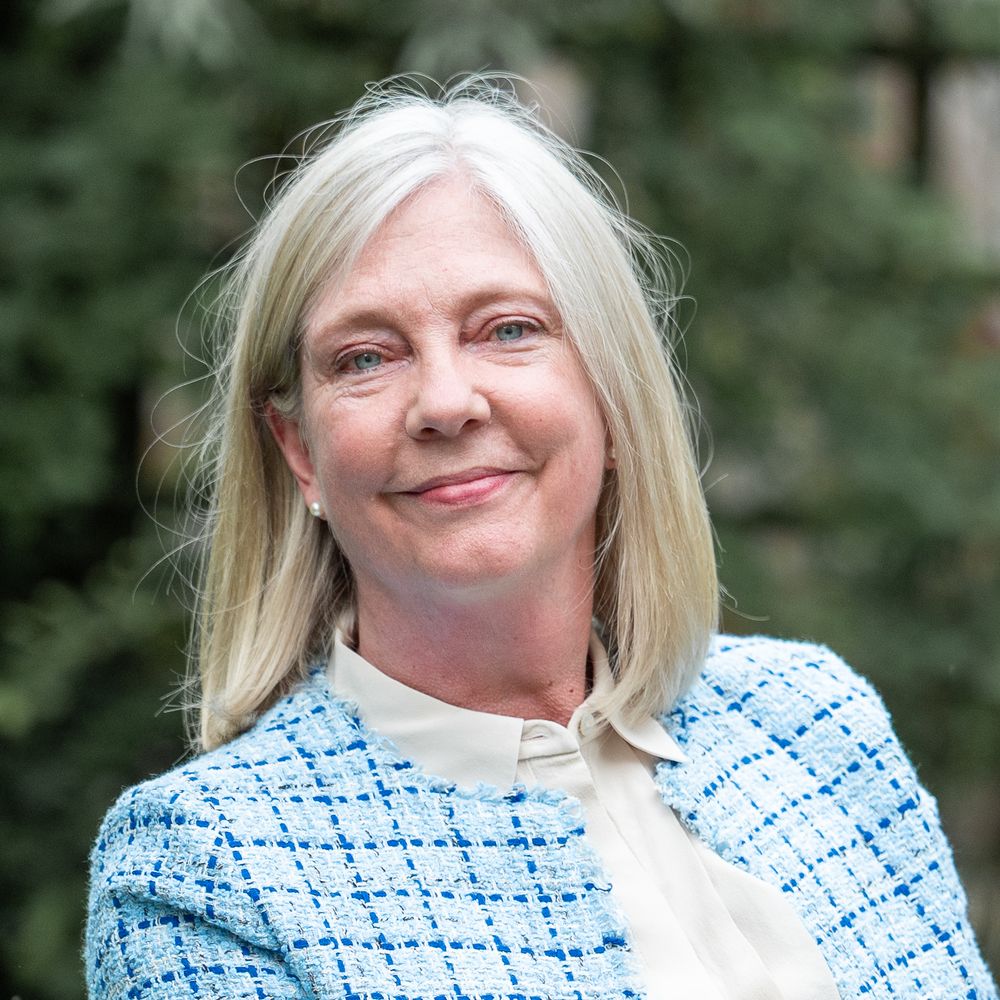 Someone wearing a light blue and white patterned jacket is smiling warmly against an outdoor background.