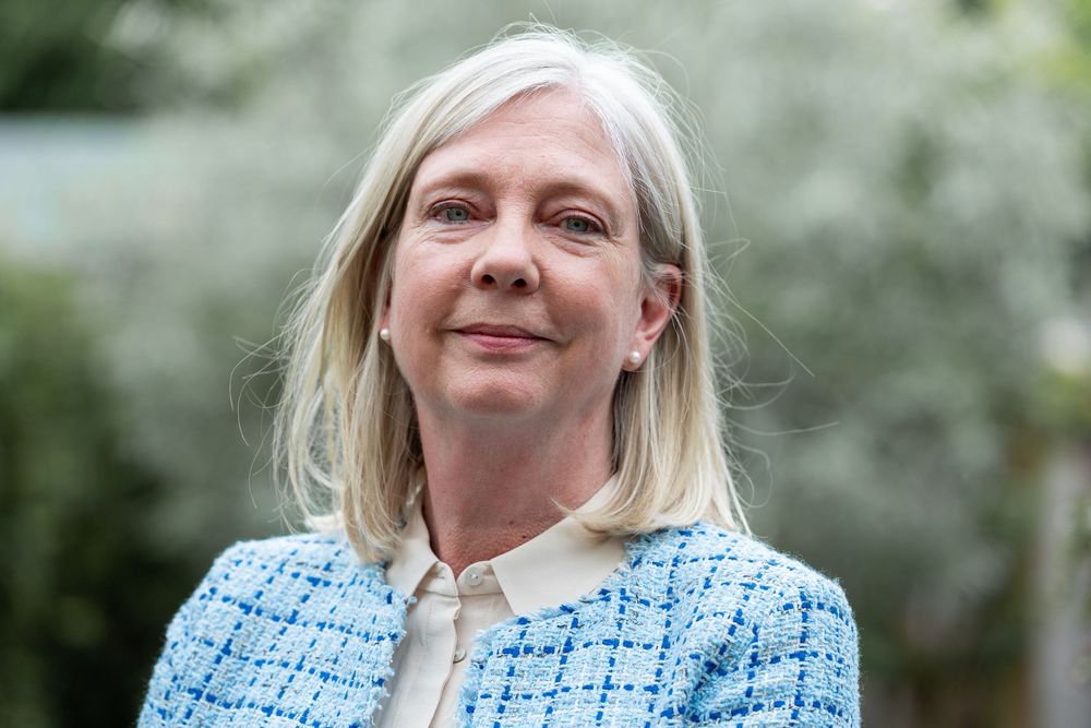 Professional portrait of a person with blonde hair wearing a light blue tweed jacket against a blurred outdoor background.