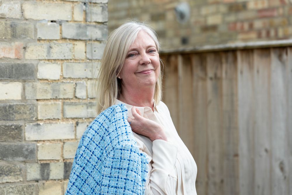 A person in a white jacket and blue plaid dress smiles while leaning against a rustic brick wall outdoors.